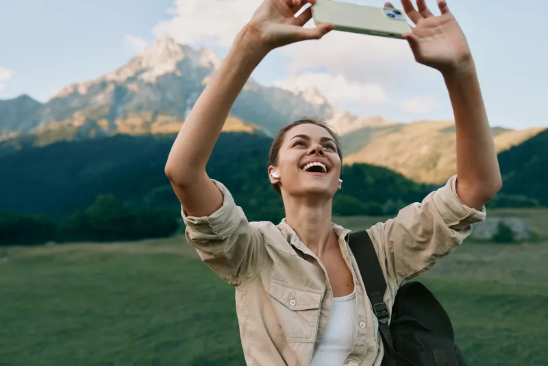 Smiling woman hiking outdoors taking a selfie against a mountainous landscape, wearing a casual beige jacket. Concept of travel, exploration, and healthy living. Natural light creates an energetic