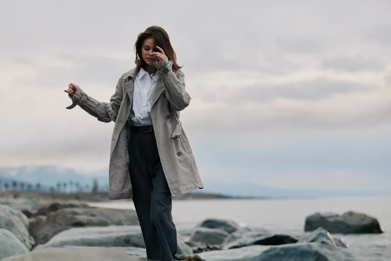 Young woman wearing a coat walks on rocky beach near the ocean under cloudy sky. Casual fashion portrait with natural landscape background and calm atmosphere.