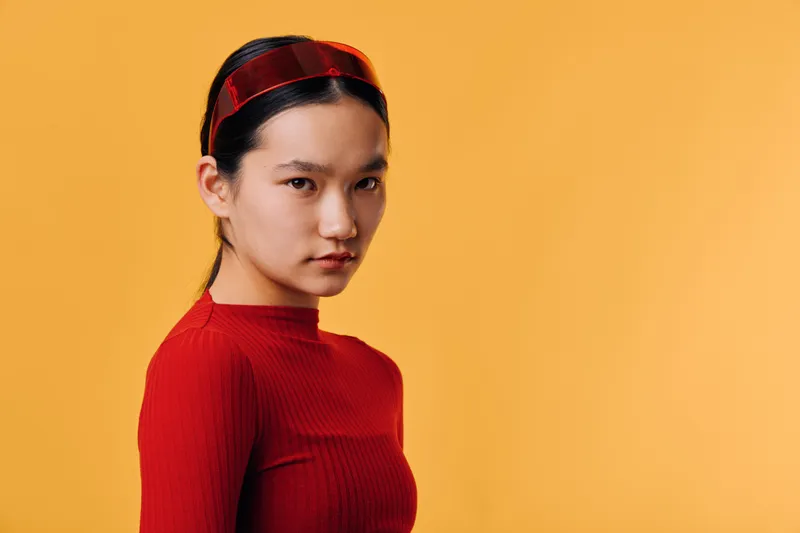Portrait of young Asian woman in red sweater and matching headband looking at camera on yellow background. Female model with serious expression and simple studio lighting for fashion or beauty.