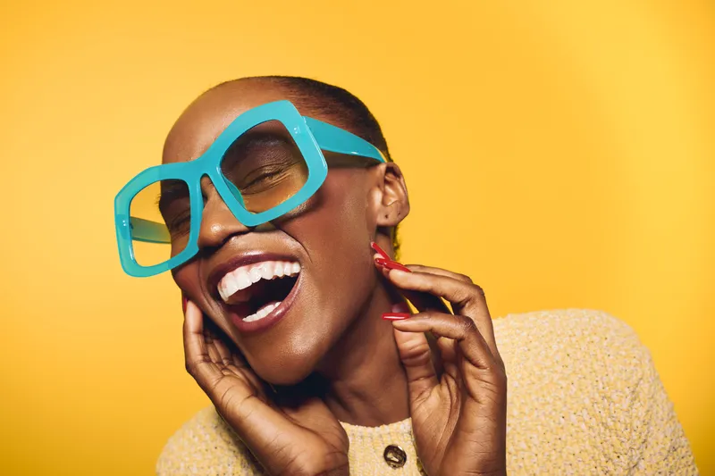 Joyful dark skinned African American woman wearing large blue sunglasses laughs with eyes closed against yellow background. Concept of happiness, fashion, beauty, and summer style.