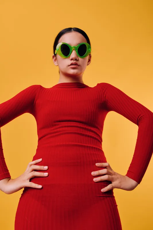 Woman in red dress with green round sunglasses posing confidently with hands on hips against yellow background. Fashion, style and bold colors in studio portrait of confident model.