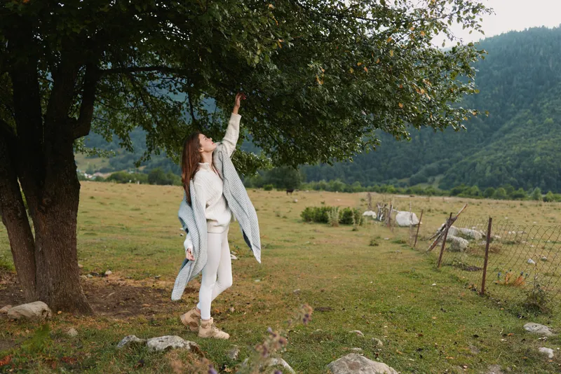 Woman in a white dress and jacket reaches toward a tree in a rural field, enjoying nature and outdoor life. A peaceful, sunlit moment with grazing cows nearby.