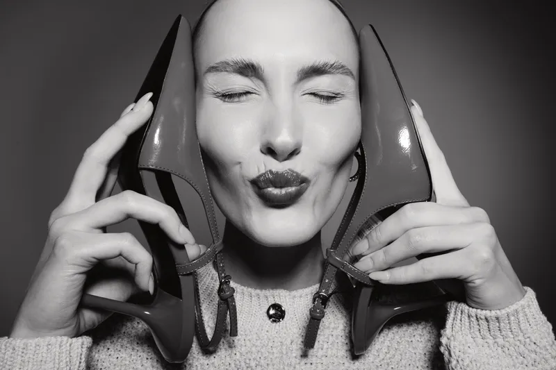 Close-up of a smiling white woman with fair skin holding red high heels near her face. Concept of fashion, footwear, joy, and female beauty in black and white photography.