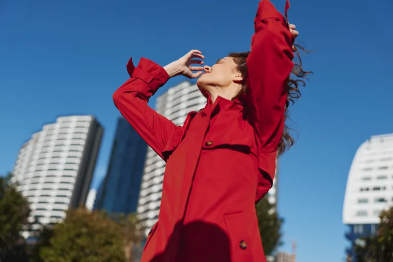Young woman in a bold red coat stands in an urban plaza, arms raised toward the sky as sunlight highlights her confident pose beside modern high rise buildings.