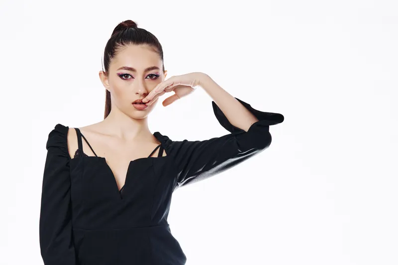 Young woman with sleek hairstyle and creative black outfit poses with fingers near face. Studio shot on white background. Concept for beauty, fashion, and high-end editorial.