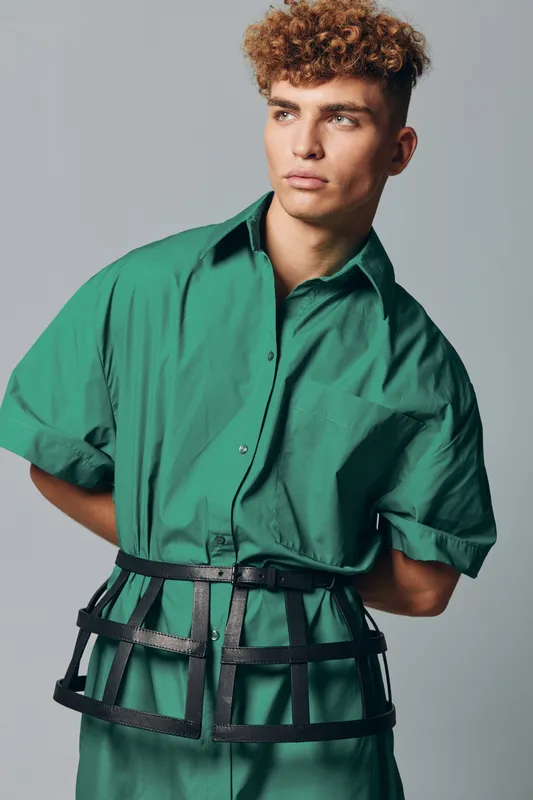 Young man with curly hair wearing green shirt and black leather cage belt posing against gray background in studio. Fashion model with serious expression and stylish outfit for editorial.
