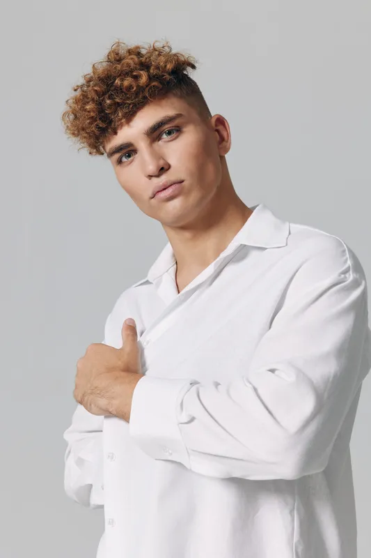 Young man with curly hair wearing white shirt poses confidently against gray background. Closeup portrait showing casual style, expression, and modern hairstyle in studio setting.