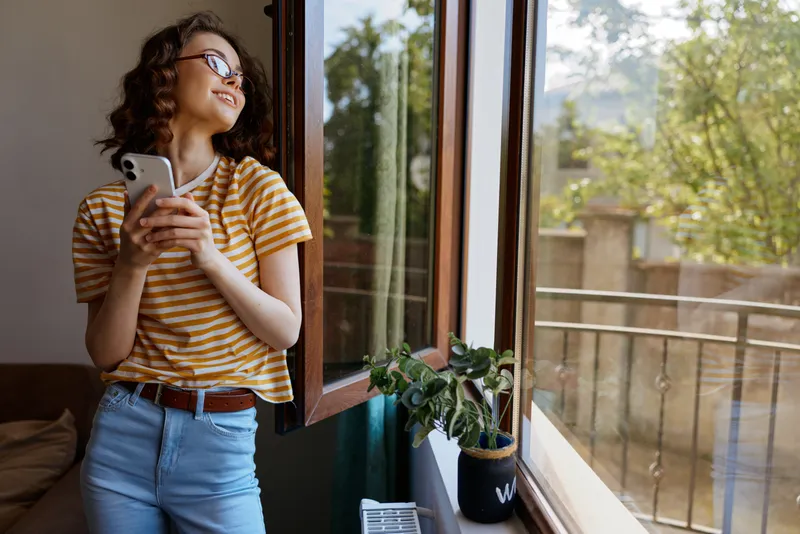 Woman stands by an open window in a striped t shirt, holding a smartphone, enjoying natural daylight at home with a potted plant nearby.