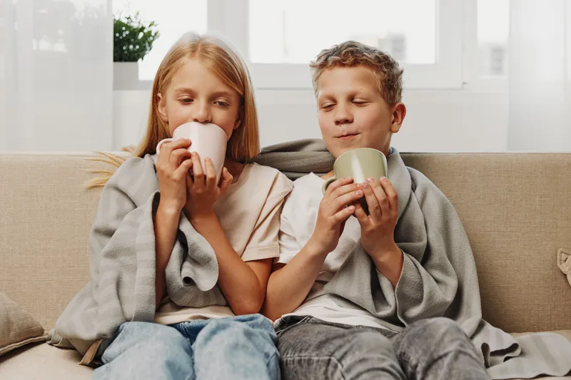 Two cozy children wrapped in blankets drink hot beverages on a sofa, enjoying a warm moment indoors with soft natural light and relaxed expressions.