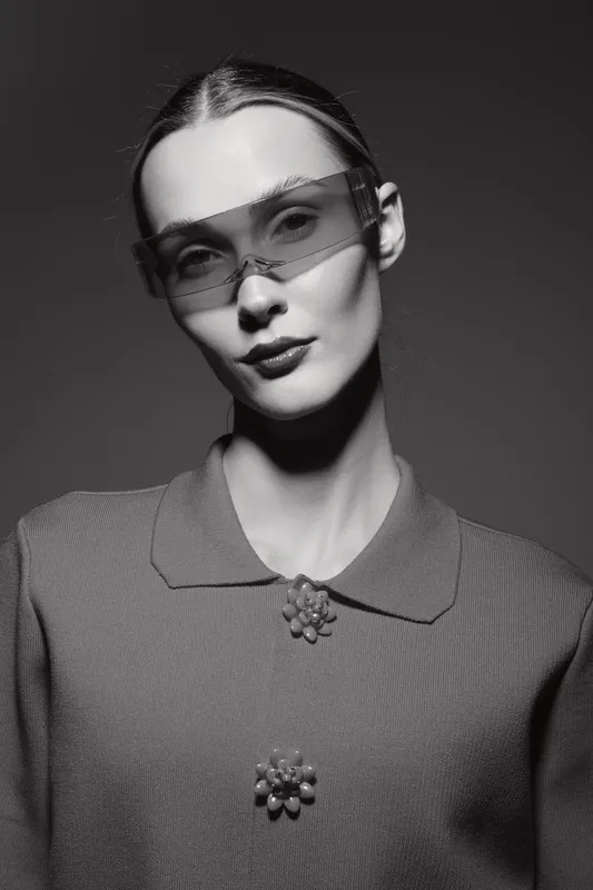 Portrait of a young Caucasian woman with pale skin wearing oversized sunglasses and a textured coat with floral buttons. Studio lighting highlights her sharp features.