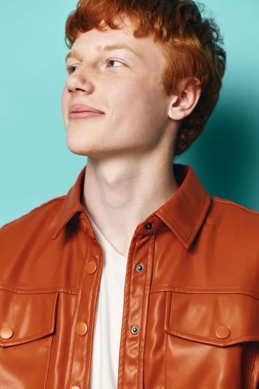 Young man with red hair wearing a stylish brown leather jacket and white shirt looks confidently to the side against a blue background with relaxed expression.