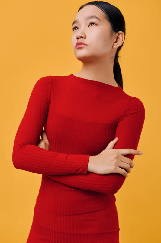 Young woman in red long sleeve sweater stands with crossed arms looking confident and thoughtful against orange background. Fashion portrait with casual style and minimalistic colors.