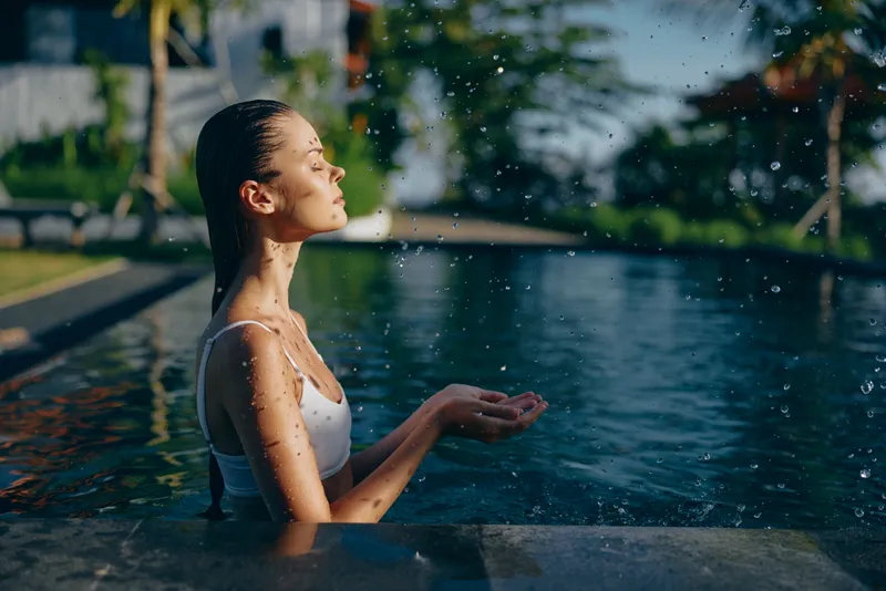 Young woman in a light swimsuit relaxes in an outdoor pool with eyes closed as water droplets glisten under bright sun, creating a calm summer moment