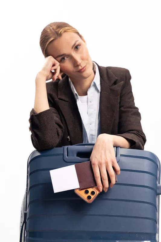 Thoughtful young woman in business attire rests head on hand while leaning on large suitcase holding passport and phone. Concept of travel, waiting, and contemplation on white background.