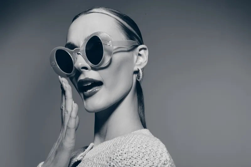 Stylish white woman with pale skin wears large round sunglasses and knitted sweater, posing with hand near face in studio. Fashion and retro concept.