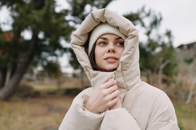 Woman wearing a beige parka with hood in an outdoor setting, winter mood with soft natural light, calm expression, and a quiet landscape behind.