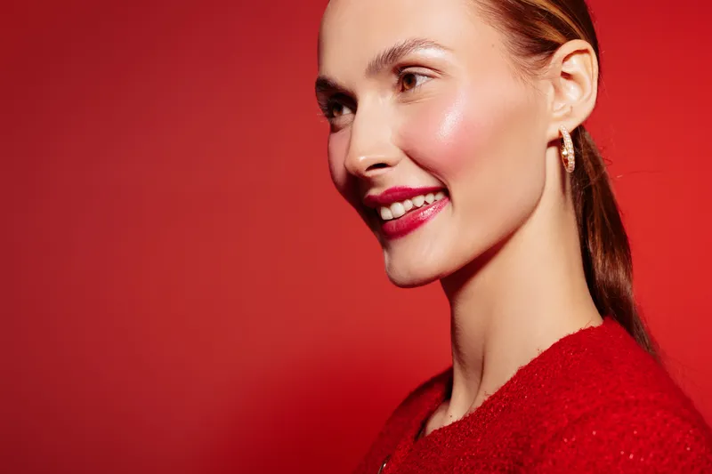 Smiling woman with red lipstick and earrings poses against red background. Close-up portrait of happy female model with natural makeup and sleek hair. Concept of beauty and fashion.