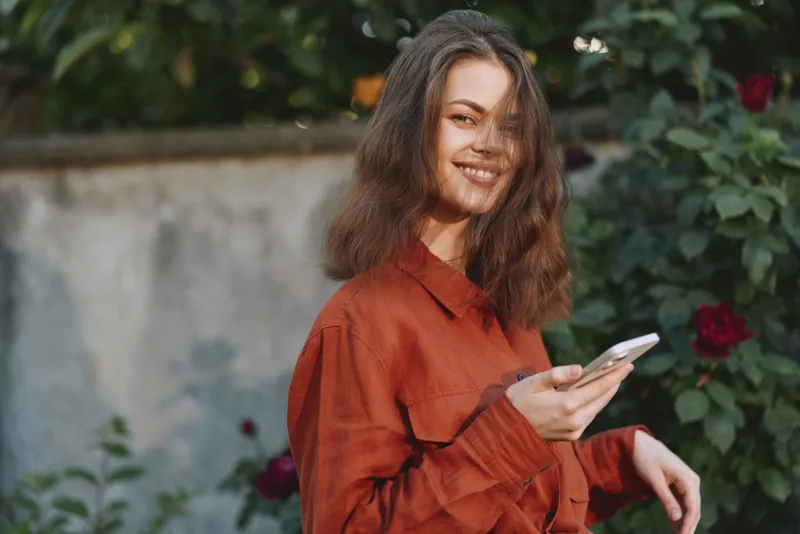 Young woman wearing a rust shirt smiles while checking her phone in a sunlit garden, enjoying a casual outdoor moment with a gentle breeze and blooming plants nearby