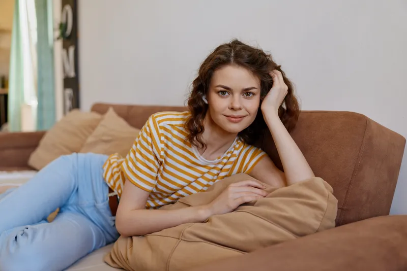 A woman relaxes on a sofa at home, wearing striped tshirt and jeans, leaning on a cushion with a gentle smile. Subtle lifestyle moment showcasing comfort and casual living.