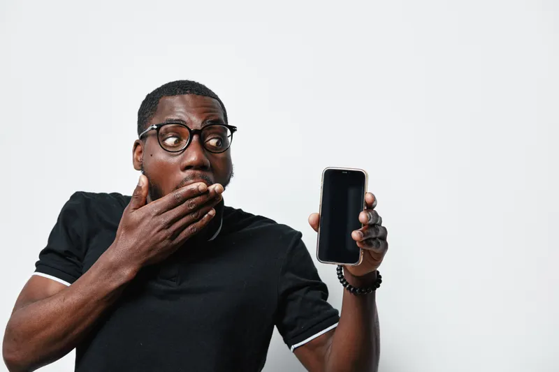 African American man with dark skin and glasses showing smartphone with blank screen and covering mouth in surprise or shock on white background. Casual clothing and expression.
