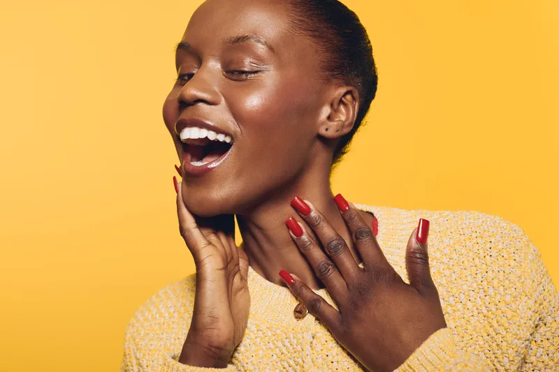 Happy dark skinned African woman with red nails laughs joyfully, wearing yellow sweater on yellow background. Concept of beauty, happiness, and self confidence in studio portrait.
