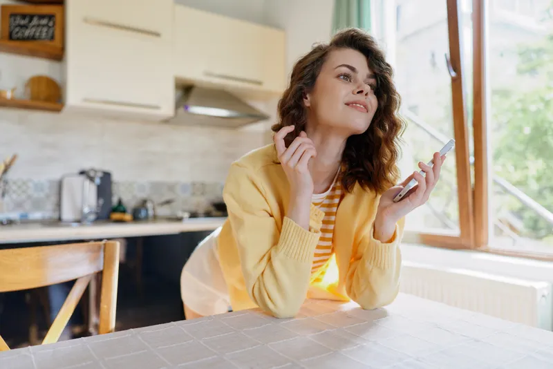 A young woman wearing a yellow cardigan sits at a kitchen table, grooming her hair and gazing toward the window with a relaxed expression.
