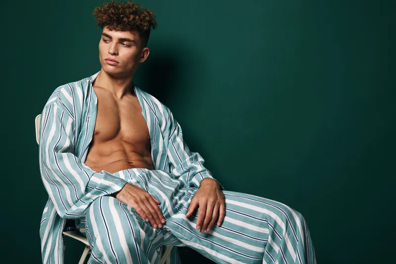 Young man with curly hair seated wearing striped pajama set without shirt. Male model in casual pose with serious expression against dark green background for fashion portrait.