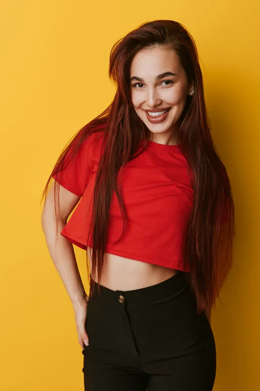 Young woman with long hair wearing red crop top and black pants smiling confidently on yellow background. Portrait of happy female model posing with hand in pocket in studio.