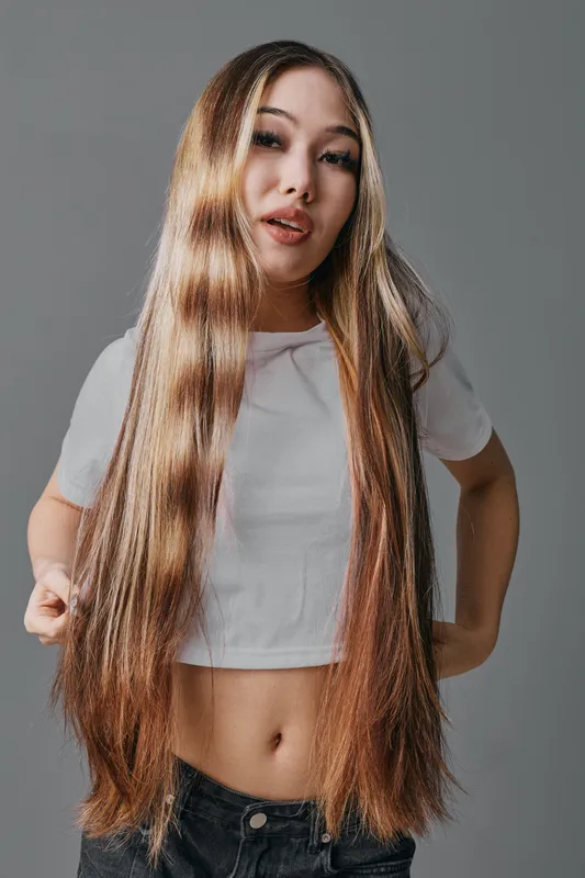 Young woman with long straight hair posing in casual white crop top and dark pants, looking confident and stylish against a neutral gray background indoors.
