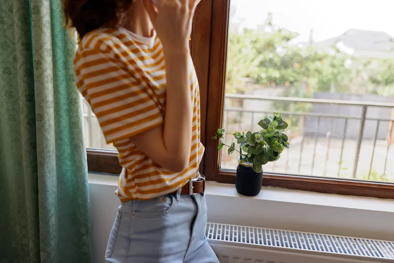 A woman stands by a sunny window, resting her chin on her hand, wearing an orange striped shirt and light shorts. A calm, intimate home moment focused on daily life.