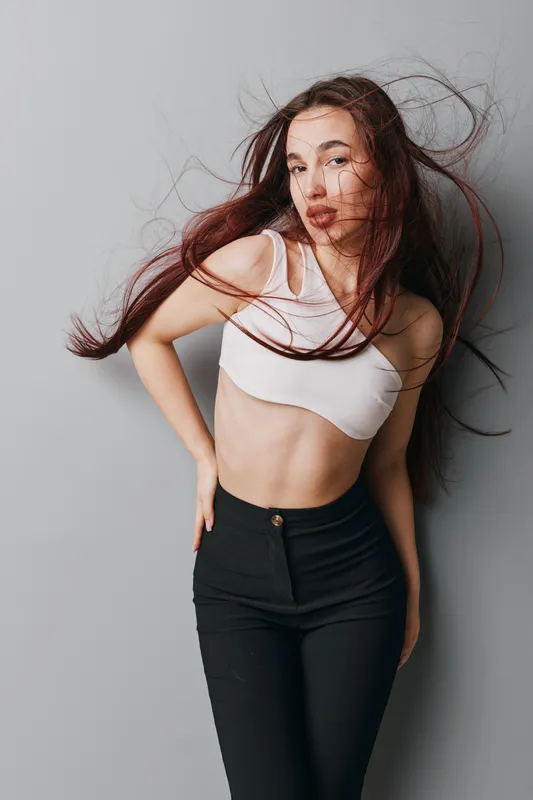 Young woman with long hair wearing white crop top and black pants posing against gray wall. Hair is flowing with movement, casual fashion style with confident expression and posture.