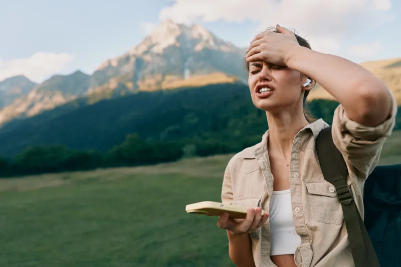 Woman traveler in a scenic field with mountains, holding a book, shielding eyes from sun, casual outfit and backpack, exploring nature and freedom