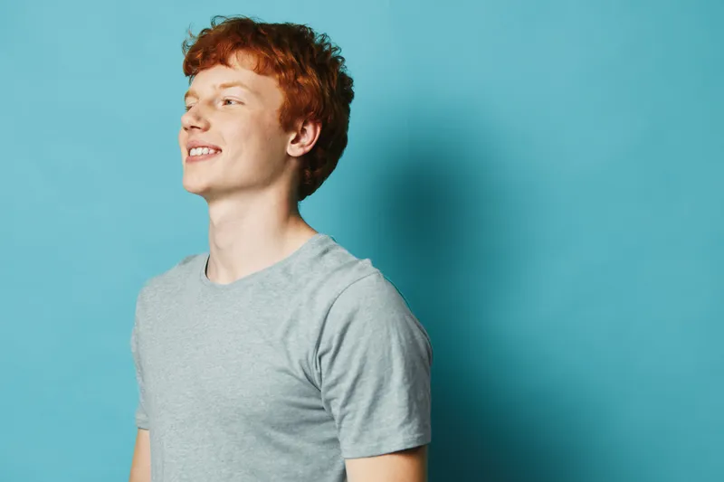 Smiling young man with red hair wearing gray shirt standing against blue background looking aside in casual style portrait with natural light and relaxed mood.