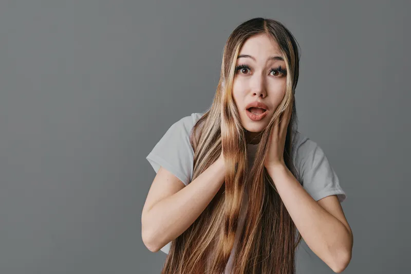 Young woman with long hair and expressive face showing surprise or shock on neutral gray background. Female model with emotions, casual wear and braided hairstyle closeup portrait.