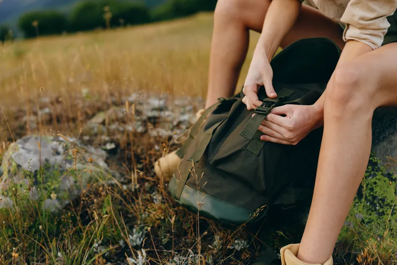 Young woman with short hair sits on grass in a natural field while opening a green backpack, enjoying a peaceful outdoor moment during a casual lifestyle hike.