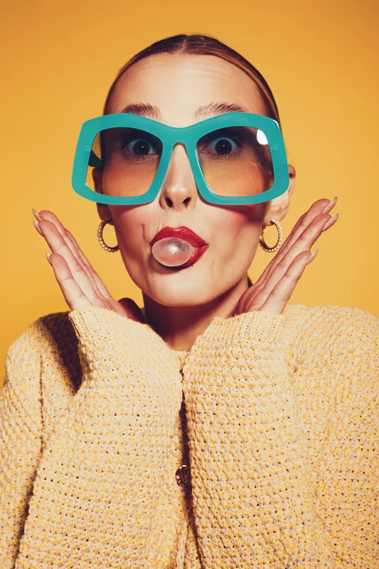 Young woman with oversized turquoise glasses blows a bubble gum bubble, wearing a yellow sweater and gold hoop earrings. Bright yellow background creates a fun and playful mood.