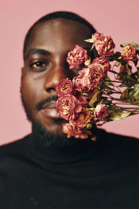 African man with dark skin holding dried pink roses close to face in creative conceptual visual style on pink background. Portrait with flowers and artistic expression in studio.