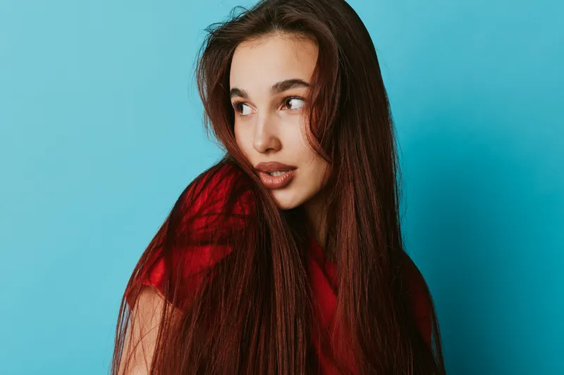 Young woman with long brown hair wearing red shirt looking sideways on blue background. Portrait of thoughtful female with natural makeup and casual style in studio lighting.