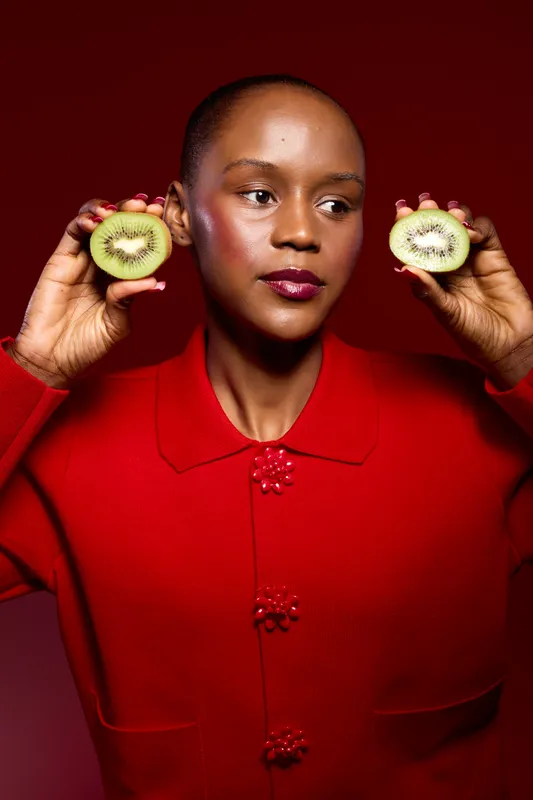 African American woman with dark skin holds two halves of green kiwi fruit near her face wearing a red sweater. Studio portrait on red background with fresh fruit and healthy lifestyle concept.