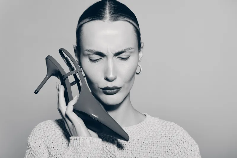 Young white woman with pale skin wearing a knitted sweater holds a high heel shoe beside her face with eyes closed. Concept of discomfort, fashion accessory, and unusual portrait.