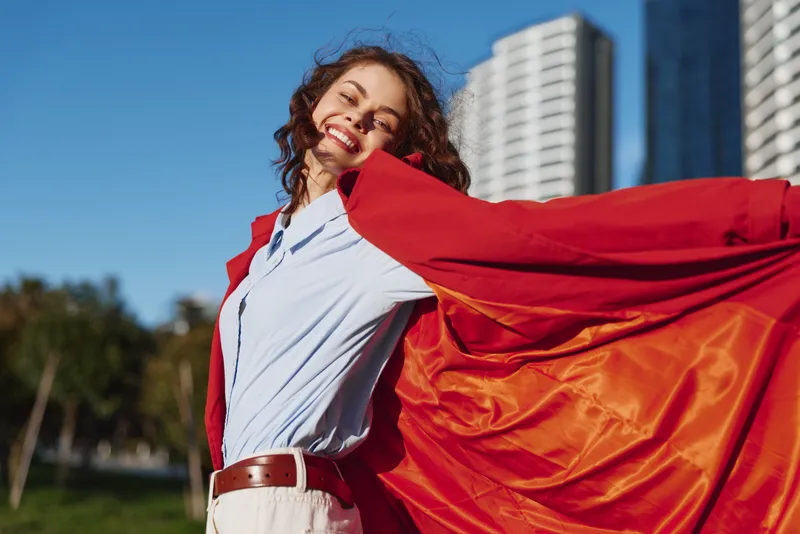 Young woman with curly hair smiles joyfully while holding a flowing red fabric behind her in an urban park on a sunny day.