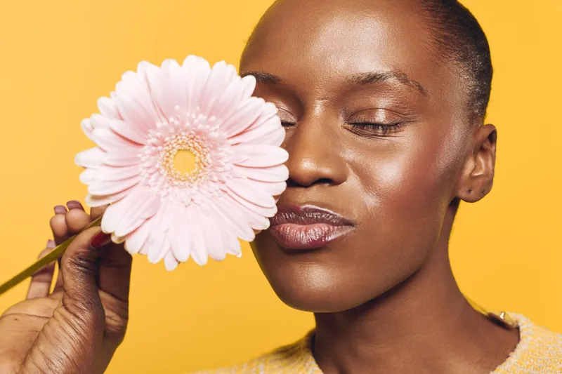 Close-up of a beautiful African woman with dark skin holding a pink gerbera flower near her face. She has closed eyes and smooth skin, isolated on a bright yellow background. Concept of beauty and