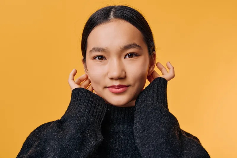 Young woman wearing black knitted sweater adjusts hair while looking at camera on yellow background. Portrait of confident female with natural makeup and casual outfit in studio lighting.
