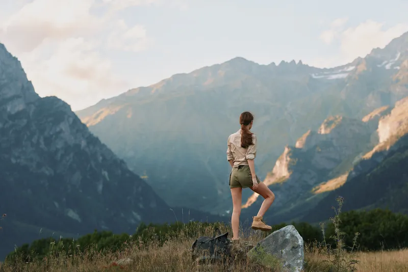Young woman stands on rock in mountain landscape wearing casual summer clothes looks at distant peaks enjoying nature and outdoor adventure during sunny day.
