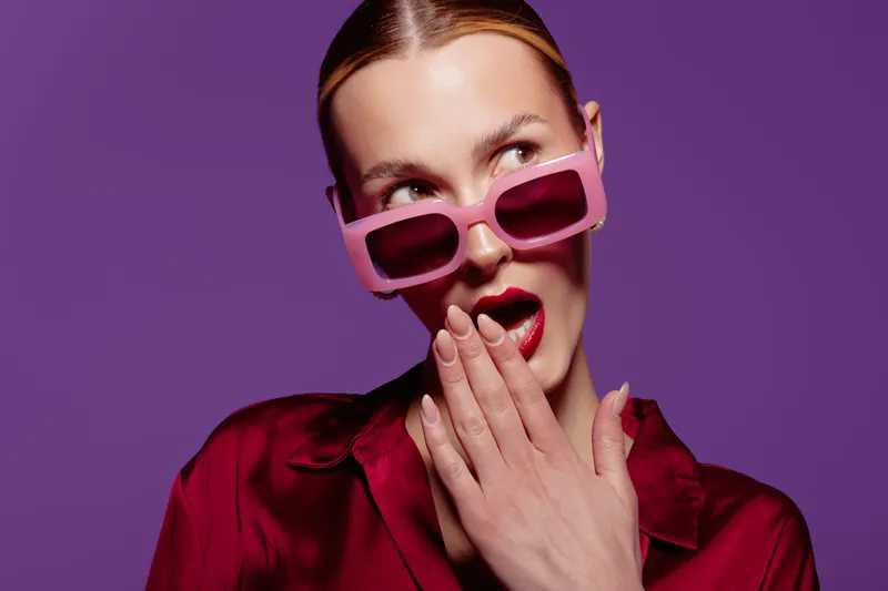 Surprised woman in pink sunglasses and red satin shirt covers mouth with hand on purple background. Concept of fashion, beauty, expression, and modern style portrait in studio lighting.