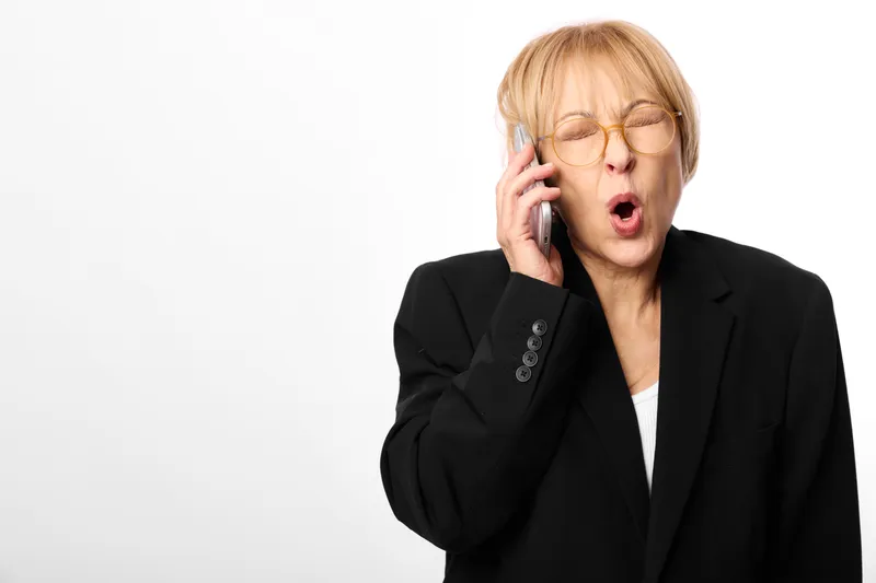 Businesswoman with a phone to her ear, eyes closed, showing surprise and stress. Studio portrait on a plain white background with soft light, during a call today.