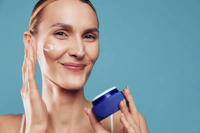 A smiling woman applies white face cream on her cheek while holding a blue jar against a plain blue background. Concept for skincare, beauty routine, and healthy skin care.