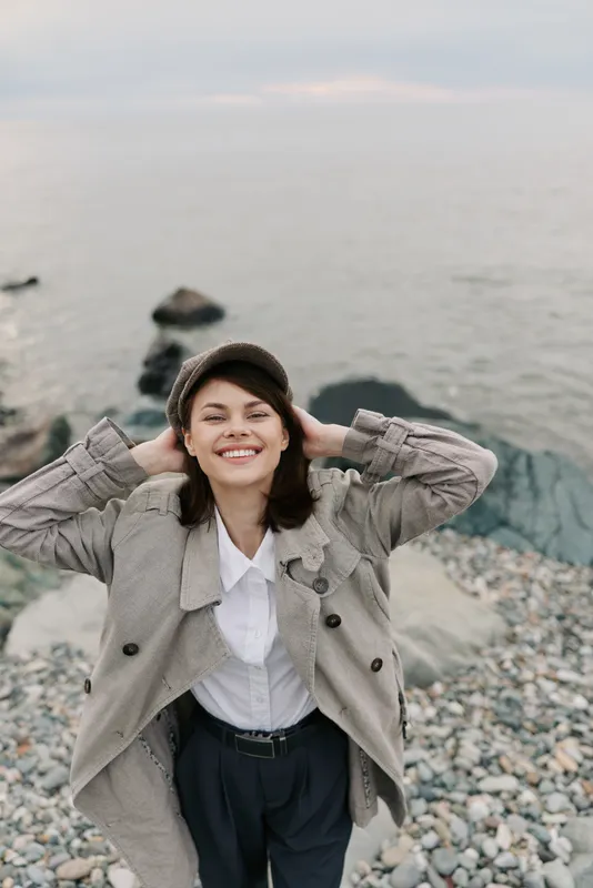 Smiling young woman in checkered coat and hat stands on rocky beach near calm sea, enjoying fresh air and peaceful moment outdoors in casual autumn outfit.