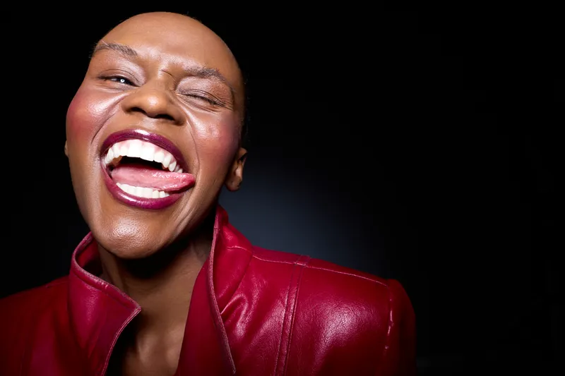 Happy African woman with dark skin wearing a red leather jacket winks and smiles with tongue out. Portrait on black background. Concept for joy, confidence, and female empowerment.