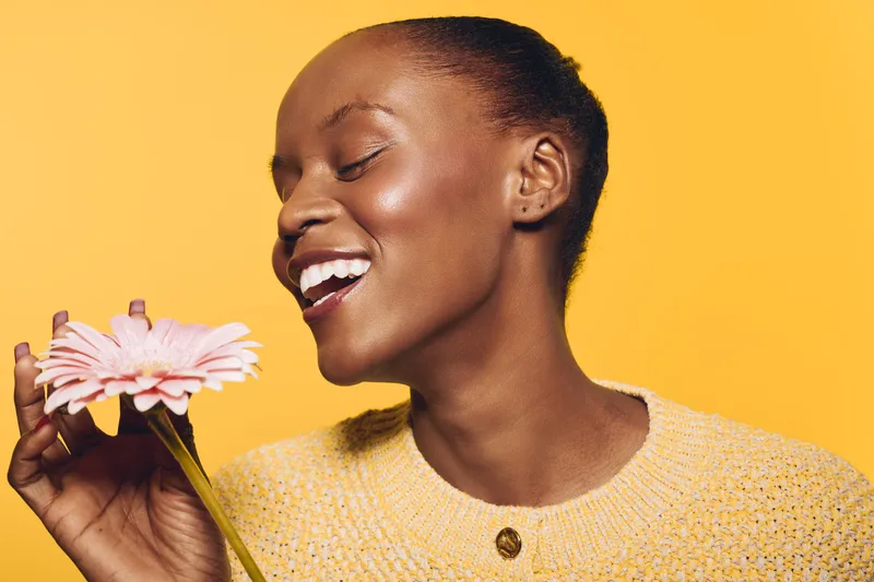 Happy smiling black woman with dark skin holds pink flower, wears yellow sweater on yellow background. Concept of natural beauty, joy, and floral wellness for African ethnicity.
